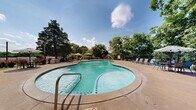 This outdoor aquatic amenity space centers around a generously sized freeform swimming pool with clear turquoise water, accented by multiple decorative water fountains that add both visual appeal and a relaxing ambiance. The pool features a gradual entry point and is framed by a spacious concrete sun deck that comfortably accommodates rows of lounge chairs and umbrellas, ideal for relaxing in the sun. A dedicated shaded seating and dining area with patio furniture and umbrellas provides a comfortable retreat from direct sunlight. The pool area is fully enclosed by a sturdy wrought iron fence, ensuring safety and privacy, with clearly posted pool rules and notices. Mature trees surround the perimeter, offering natural shade and a verdant backdrop that enhances the resort-like atmosphere. Vibrant flowering plants and manicured greenery line the fence line, adding color and charm to the setting. An adjacent multi-story brick building with white-trimmed balconies overlooks the pool, providing easy access to the amenity. The overall design creates an inviting, well-maintained outdoor retreat suited for warm-weather relaxation and recreation.