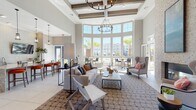 This single-floor amenity space is designed with an open, airy layout that immediately draws attention to its soaring double-height ceilings accented by exposed wood beams and a striking coffered ceiling detail. A dramatic curved wall of floor-to-ceiling windows floods the main lounge area with abundant natural light while framing views of a resort-style pool, palm trees, and the surrounding community beyond.

The main lounge is anchored by a generous seating arrangement of neutral-toned sofas and wingback chairs centered around a sleek coffee table, all set atop a patterned area rug over large-format tile flooring. An iron chandelier adds an elegant focal point overhead. A full-height stacked stone accent wall houses a linear gas fireplace, creating a sophisticated gathering spot flanked by upholstered seating.

An adjacent kitchen and bar area features a substantial granite-topped island with seating, white cabinetry, a textured tile backsplash, and a wall-mounted flat-screen television—ideal for social gatherings. Glass-paneled folding doors connect the main hall to a bright sunroom-style corridor lined with windows, extending the sense of openness throughout. High-top bistro tables positioned along the window wall provide intimate seating with direct outdoor views.
