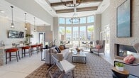 This single-floor amenity space is designed with an open, airy layout that immediately draws attention to its soaring double-height ceilings accented by exposed wood beams and a striking coffered ceiling detail. A dramatic curved wall of floor-to-ceiling windows floods the main lounge area with abundant natural light while framing views of a resort-style pool, palm trees, and the surrounding community beyond.

The main lounge is anchored by a generous seating arrangement of neutral-toned sofas and wingback chairs centered around a sleek coffee table, all set atop a patterned area rug over large-format tile flooring. An iron chandelier adds an elegant focal point overhead. A full-height stacked stone accent wall houses a linear gas fireplace, creating a sophisticated gathering spot flanked by upholstered seating.

An adjacent kitchen and bar area features a substantial granite-topped island with seating, white cabinetry, a textured tile backsplash, and a wall-mounted flat-screen television—ideal for social gatherings. Glass-paneled folding doors connect the main hall to a bright sunroom-style corridor lined with windows, extending the sense of openness throughout. High-top bistro tables positioned along the window wall provide intimate seating with direct outdoor views.