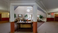 Located at 1480 Park St in White Bear Lake, Minnesota, this single-floor community space offers a warm and inviting atmosphere designed for comfort and gathering. The centerpiece of the space is a striking rotunda-style lobby featuring soaring vaulted ceilings adorned with decorative floral garlands, terracotta accent walls, and an ornate patterned carpet. An elegant chandelier anchors the space, which is furnished with a loveseat and coffee table, creating a relaxed yet refined common area. Adjacent to the lobby, a welcoming lounge area features additional seating, framed wall art, decorative mirrors, and lush indoor plants, all bathed in natural light from large windows. A dedicated entry foyer connects to the outdoors through full-height glass doors, offering views of mature trees and a landscaped exterior. This bright, tile-floored sunroom includes a glass-top dining table with wrought-iron chairs, ideal for casual conversation or informal gatherings. Crown molding and textured ceilings add architectural character throughout. A carpeted hallway with warm wall sconces and terracotta wainscoting ties the various areas together, providing a cohesive and navigable layout. The overall ambiance is hospitable, well-appointed, and thoughtfully designed for community living.