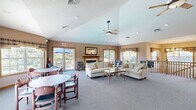 Located at 454 Crestmont Ct. in Danbury Ct., Ohio, this single-floor community clubhouse offers a well-appointed and inviting gathering space designed for comfort and functionality. The entrance welcomes visitors through glass doors into a tiled hallway, accented by textured wallpaper and a console table, setting a warm and approachable tone throughout.

The expansive main lounge is the centerpiece of the space, featuring soaring vaulted ceilings and a ceiling fan that enhance the sense of openness and airiness. A classic wood-mantel fireplace anchors one wall, flanked by comfortable sofas and a glass coffee table, creating an ideal setting for relaxed gatherings. Plush carpeting and neutral warm tones contribute to the welcoming ambiance. Multiple windows and sliding glass doors flood the room with natural light and provide direct access to an outdoor deck, offering pleasant views of the surrounding greenery.

A dedicated dining area with round tables and chairs is situated adjacent to the lounge, also benefiting from abundant natural light through large windows. The compact kitchen features light wood cabinetry, laminate countertops, a stainless steel sink, and tile flooring, providing practical support for events and gatherings. A staircase connects to additional levels, and a bathroom is also included. The overall layout is thoughtfully designed to serve as a functional and comfortable community hub.