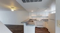 This two-story space presents a functional and clean layout highlighted by an open-concept kitchen that flows seamlessly into the adjoining living area. The kitchen is equipped with white cabinetry offering generous storage, a white electric range, and under-cabinet ventilation. A peninsula with a grey laminate countertop and a double-basin sink serves as a natural divider between the kitchen and living space, providing additional workspace and casual seating potential. Recessed and flush-mounted lighting fixtures illuminate the area evenly, complementing the bright white walls and vaulted ceiling detail that adds a sense of vertical openness to the space. Dark hardwood-style flooring runs throughout, providing a warm contrast to the lighter upper elements. The white tones and clean lines throughout the kitchen create a neutral, move-in-ready aesthetic. The second floor adds additional living or sleeping space, expanding the overall footprint of the unit. The combination of practical storage solutions, open sightlines, and durable finishes makes this a well-appointed space suited for everyday living.