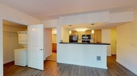 Located at 910 8th Avenue in the heart of Seattle, Unit 405 is a thoughtfully designed single-floor residence that balances functionality with comfortable urban living. The open-concept layout connects the living area to a peninsula-style kitchen, creating a cohesive and efficient use of space. Rich hardwood-style flooring runs throughout the main living areas, adding warmth and continuity to the design.

The kitchen is well-equipped with light maple cabinetry, dark stone-look countertops, and a full suite of black appliances including a refrigerator, electric range, built-in microwave, and ample cabinet storage. Pendant lighting above the peninsula adds an elegant finishing touch.

The living area is bright and welcoming, featuring a large floor-to-ceiling window wall that floods the space with natural light and provides a lush, tree-lined outdoor view. A door leads directly to a private outdoor area, ideal for enjoying Seattle's greenery.

The bathroom is generously sized, offering an extended vanity with light maple cabinetry, a broad countertop, a large mirror, and a combination tub and shower with white tile surround. Additional storage is conveniently integrated along the vanity wall.

A dedicated walk-in closet provides substantial storage with built-in wire shelving and hanging rods. The unit also includes a washer and dryer, as visible in the floor plan, adding everyday convenience to this practical and comfortable Seattle residence.