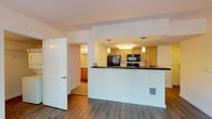 Located at 910 8th Avenue in the heart of Seattle, Unit 405 is a thoughtfully designed single-floor residence that balances functionality with comfortable urban living. The open-concept layout connects the living area to a peninsula-style kitchen, creating a cohesive and efficient use of space. Rich hardwood-style flooring runs throughout the main living areas, adding warmth and continuity to the design.

The kitchen is well-equipped with light maple cabinetry, dark stone-look countertops, and a full suite of black appliances including a refrigerator, electric range, built-in microwave, and ample cabinet storage. Pendant lighting above the peninsula adds an elegant finishing touch.

The living area is bright and welcoming, featuring a large floor-to-ceiling window wall that floods the space with natural light and provides a lush, tree-lined outdoor view. A door leads directly to a private outdoor area, ideal for enjoying Seattle's greenery.

The bathroom is generously sized, offering an extended vanity with light maple cabinetry, a broad countertop, a large mirror, and a combination tub and shower with white tile surround. Additional storage is conveniently integrated along the vanity wall.

A dedicated walk-in closet provides substantial storage with built-in wire shelving and hanging rods. The unit also includes a washer and dryer, as visible in the floor plan, adding everyday convenience to this practical and comfortable Seattle residence.