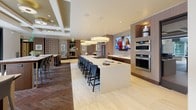 This elegantly designed clubhouse spans a single floor and offers a refined blend of modern aesthetics and functional communal spaces. The open-concept layout centers around a striking oversized kitchen island topped with a pristine white quartz surface, surrounded by quilted leather bar seating. The kitchen is fully equipped with stainless steel appliances, including a gas cooktop, double wall ovens, and a full-size refrigerator, all framed by warm wood cabinetry and a shimmering tile backsplash. Statement drum pendant lights hang above, adding a polished finishing touch. Adjacent to the kitchen, a spacious lounge area features plush sectional seating and large folding glass doors that open directly onto an outdoor terrace, creating a seamless indoor-outdoor flow with views of palm trees and the surrounding cityscape. A decorative lattice screen divides the bar seating area from the lounge, adding architectural interest while maintaining an open feel. Hardwood herringbone flooring transitions throughout the social areas, grounding the space with warmth and texture. A thoughtfully appointed restroom features a marble vanity, wood-paneled walls, and an intricate mosaic tile accent wall. Curated artwork and refined furnishings complete the upscale ambiance of this impressive community clubhouse.