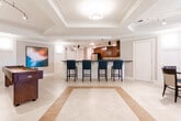 Located at 140 Mayhill Street in Saddle Brook, NJ, this single-floor amenity space is thoughtfully designed for socializing and relaxation. The open main area features elegant cream-toned tile flooring with a decorative inlaid border, setting a refined tone throughout. A full-length bar with dark granite countertops and navy blue upholstered bar stools serves as a central gathering point, complemented by rich walnut cabinetry and pendant lighting overhead. Adjacent to the bar, a well-equipped kitchen showcases warm wood cabinetry, dark granite countertops, a multicolor mosaic tile backsplash, stainless steel appliances including a full-size refrigerator and microwave, and a stainless steel sink — ideal for catering and entertaining. A dedicated lounge area offers plush seating arranged around a glass-top coffee table, anchored by a built-in walnut entertainment wall unit with a large flat-screen TV and display shelving. Track lighting and a decorative drum pendant light enhance the cozy ambiance. The dining area accommodates multiple groups at glass-top tables with upholstered chairs, bathed in natural light from large windows with garden views. Bold abstract artwork adds a contemporary artistic touch throughout the space.