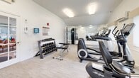 Located at 140 Plains Rd in Windham, Connecticut, this fitness center offers a clean, well-organized workout facility contained within a single floor. The space features vaulted ceilings that lend an open, airy feel, complemented by bright white walls and light wood-style flooring that create a modern, welcoming atmosphere.

The facility is comprehensively equipped to support a full range of workout routines. Cardio equipment includes elliptical trainers, a recumbent bike, a stationary lifecycle bike, and a treadmill. For strength training, the center provides a Precor functional trainer cable machine, an adjustable weight bench, and a rack of dumbbells spanning a variety of weights. Large wall-mounted mirrors enhance the sense of space and allow for proper form checks during exercise.

Natural light fills portions of the room through windows fitted with vertical blinds, while flush-mounted overhead lighting ensures consistent illumination throughout. The space also includes a wall-mounted AED unit and posted fitness center rules for user safety and guidance.

A dedicated computer workstation area is situated within the room, offering a convenient space for administrative use or digital fitness tracking. The overall environment is clean, bright, and purposefully arranged to maximize usability and comfort for all fitness levels.