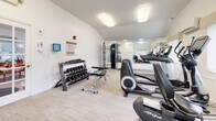Located at 140 Plains Rd in Windham, Connecticut, this fitness center offers a clean, well-organized workout facility contained within a single floor. The space features vaulted ceilings that lend an open, airy feel, complemented by bright white walls and light wood-style flooring that create a modern, welcoming atmosphere.

The facility is comprehensively equipped to support a full range of workout routines. Cardio equipment includes elliptical trainers, a recumbent bike, a stationary lifecycle bike, and a treadmill. For strength training, the center provides a Precor functional trainer cable machine, an adjustable weight bench, and a rack of dumbbells spanning a variety of weights. Large wall-mounted mirrors enhance the sense of space and allow for proper form checks during exercise.

Natural light fills portions of the room through windows fitted with vertical blinds, while flush-mounted overhead lighting ensures consistent illumination throughout. The space also includes a wall-mounted AED unit and posted fitness center rules for user safety and guidance.

A dedicated computer workstation area is situated within the room, offering a convenient space for administrative use or digital fitness tracking. The overall environment is clean, bright, and purposefully arranged to maximize usability and comfort for all fitness levels.