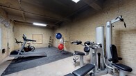 This fitness facility offers a practical and functional workout environment designed to accommodate a range of exercise needs. The space is defined by its distinctive exposed brick walls, painted in a light cream tone, lending the room an industrial character that is both raw and utilitarian. The ceiling remains open and unfinished, with fluorescent strip lighting providing consistent, even illumination throughout.

The gym floor is partially covered with rubber matting in the workout zones, offering cushioning and grip for exercise activities. A multi-station strength training machine anchors the space on one side, providing users with a variety of resistance training options. Alongside it, a dedicated bench adds additional versatility for strength-focused workouts.

Cardio is well represented with a treadmill positioned along the far wall, suitable for both walking and running training. A ball rack holds an assortment of exercise balls in various sizes, supporting stretching, balance, and core conditioning routines. A corkboard is mounted on the wall, offering a practical space for posting schedules, instructions, or fitness plans.

The layout is open and spacious, allowing for free movement between equipment stations. Overall, this gym presents a no-frills, purposeful training environment suited to a broad range of fitness activities.