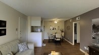 This single-floor residence showcases an open-concept layout that seamlessly connects the living, dining, and kitchen areas into one cohesive space. The living area, visible in the foreground, features a light-toned sofa with decorative accent pillows and a coffee table, set atop rich, dark hardwood-style flooring that extends throughout the main area. Warm gray accent walls contrast with crisp white trim and baseboards, creating a clean, modern aesthetic.

The dining area is centrally positioned and accommodates a round glass-top table surrounded by white and dark-framed chairs, complemented by wall art and decorative accents that add personality to the space. A ceiling fan overhead provides both practical ventilation and ambient lighting to this zone.

A pass-through opening connects the main living area to the kitchen, which features white cabinetry and a countertop bar ledge — an efficient design that maintains visual openness while defining the cooking space. A hallway visible on the right side of the space leads to additional rooms. The textured ceiling and consistent flooring throughout contribute to a unified and well-maintained interior.