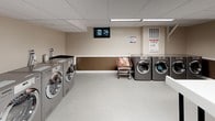 This shared laundry and utility facility at 2901 Connecticut Ave in Washington, DC, is a well-organized, single-floor space designed to serve the practical needs of its users. The room features a suspended drop ceiling with integrated fluorescent lighting panels that provide bright, even illumination throughout. The walls are finished in a two-tone scheme — neutral beige on the upper portion and a warm brown accent along the lower half — creating a clean and functional environment.

The laundry area is well-equipped with multiple stainless-steel front-loading washers and dryers arranged along the walls, offering ample capacity for shared use. A utility sink is also present, adding further convenience. Laundry carts are available for transporting items throughout the space. Folding tables are positioned centrally and along the walls, providing generous surface area for sorting and folding laundry.

A dedicated area includes a vending machine for laundry supplies, an informational bulletin board, and posted usage guidelines, ensuring users have everything they need on hand. The space also incorporates a waste and recycling station, with clearly labeled large bins for commingled recyclables, supporting responsible waste management. A fire safety pull station is visibly mounted on the wall, reflecting attention to safety standards. Light-toned vinyl tile flooring completes the practical, easy-to-maintain interior.