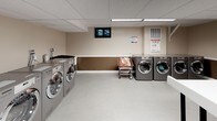 This shared laundry and utility facility at 2901 Connecticut Ave in Washington, DC, is a well-organized, single-floor space designed to serve the practical needs of its users. The room features a suspended drop ceiling with integrated fluorescent lighting panels that provide bright, even illumination throughout. The walls are finished in a two-tone scheme — neutral beige on the upper portion and a warm brown accent along the lower half — creating a clean and functional environment.

The laundry area is well-equipped with multiple stainless-steel front-loading washers and dryers arranged along the walls, offering ample capacity for shared use. A utility sink is also present, adding further convenience. Laundry carts are available for transporting items throughout the space. Folding tables are positioned centrally and along the walls, providing generous surface area for sorting and folding laundry.

A dedicated area includes a vending machine for laundry supplies, an informational bulletin board, and posted usage guidelines, ensuring users have everything they need on hand. The space also incorporates a waste and recycling station, with clearly labeled large bins for commingled recyclables, supporting responsible waste management. A fire safety pull station is visibly mounted on the wall, reflecting attention to safety standards. Light-toned vinyl tile flooring completes the practical, easy-to-maintain interior.