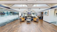 This inviting single-floor community clubhouse offers a well-appointed gathering space defined by its dramatic vaulted ceilings and striking exposed white timber truss framework. The open-concept lounge area serves as the centerpiece, featuring rich wood-look tile flooring, an elegant fireplace, a wall-mounted television, and a carefully curated mix of plush sectional sofas and accent chairs in deep purples and warm yellows. Natural light pours in abundantly through clerestory windows positioned high along the roofline, as well as through large glass entry doors that offer views of the surrounding greenery. A glass-walled fitness center runs along one side of the main lounge, keeping the space visually open while providing a dedicated workout area. Adjacent to the lounge, a leasing office enclosed in frosted glass panels offers a professional yet transparent workspace. The reception and entry area is generously proportioned, with an airy, open feel and clear sightlines throughout. A fully equipped kitchen rounds out the amenities, featuring stainless steel appliances including a range, dishwasher, and refrigerator, complemented by two-tone cabinetry in white and slate blue, a subway tile backsplash, and granite-style countertops. The overall ambiance is modern, welcoming, and thoughtfully designed for both community gatherings and everyday convenience.