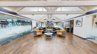 This inviting single-floor community clubhouse offers a well-appointed gathering space defined by its dramatic vaulted ceilings and striking exposed white timber truss framework. The open-concept lounge area serves as the centerpiece, featuring rich wood-look tile flooring, an elegant fireplace, a wall-mounted television, and a carefully curated mix of plush sectional sofas and accent chairs in deep purples and warm yellows. Natural light pours in abundantly through clerestory windows positioned high along the roofline, as well as through large glass entry doors that offer views of the surrounding greenery. A glass-walled fitness center runs along one side of the main lounge, keeping the space visually open while providing a dedicated workout area. Adjacent to the lounge, a leasing office enclosed in frosted glass panels offers a professional yet transparent workspace. The reception and entry area is generously proportioned, with an airy, open feel and clear sightlines throughout. A fully equipped kitchen rounds out the amenities, featuring stainless steel appliances including a range, dishwasher, and refrigerator, complemented by two-tone cabinetry in white and slate blue, a subway tile backsplash, and granite-style countertops. The overall ambiance is modern, welcoming, and thoughtfully designed for both community gatherings and everyday convenience.