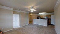 Located at 8400 Brass Mill Ln in Raleigh, NC, this single-floor residence offers a well-designed, open-concept layout that maximizes both functionality and comfort. The main living area is generously sized, finished with plush wall-to-wall carpeting and neutral-toned walls that create a warm, inviting atmosphere. Crown molding lines the ceiling throughout, adding a refined architectural detail, while a classic white ceiling fan with a light fixture provides both air circulation and ambient lighting. Decorative columns subtly define the transition between living spaces, lending a sense of structure without sacrificing the open feel. The kitchen is positioned at the rear of the living space and is accessible via a breakfast bar counter, which serves as both a prep surface and a casual dining area. The kitchen is equipped with wood-toned cabinetry, including an upper wine rack, black appliances including a refrigerator and range, and a built-in microwave. Fluorescent overhead lighting ensures the kitchen is well-lit for meal preparation. A white paneled door provides access to additional storage or utility space. The overall ambiance is clean, practical, and move-in ready, making it an ideal residence in the Raleigh area.