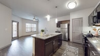 This single-floor residence offers a thoughtfully designed open-concept layout that seamlessly connects the kitchen and living areas. The kitchen serves as the centerpiece of the space, anchored by a large island with a granite countertop and double-basin sink, illuminated by stylish pendant lighting. Rich dark cabinetry lines the kitchen walls, complemented by under-cabinet lighting, black appliances including a range and microwave, and a stainless steel refrigerator. A decorative lattice detail above the cabinetry adds a distinctive architectural touch. Dark hardwood-style flooring flows throughout the main living areas, creating a warm and cohesive aesthetic, while ceiling fans in both the kitchen and living areas provide functional comfort. The living room benefits from natural light streaming through multiple windows with blinds and features direct access to an exterior door. The bedrooms are finished with plush carpeting, ceiling fans, and generous windows that invite natural light. The bathroom is clean and functional, featuring a tub-shower combination, dark vanity cabinetry with granite countertops, and wood-style flooring. Neutral wall tones throughout unify the space with a contemporary, move-in-ready appeal.