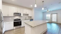 Located at Flagler Crossing in St. Augustine, FL, this well-appointed one-bedroom, one-bathroom residence offers a thoughtfully designed single-floor layout. The open-concept kitchen and living area serves as the heart of the home, showcasing a central granite-topped island with pendant lighting overhead, crisp white shaker cabinetry, a subway tile backsplash, and a full suite of stainless steel appliances including a refrigerator, range, and microwave. Light wood-style flooring runs throughout the main living areas, complementing the bright, neutral color palette. The living room benefits from natural light through a window and a glass door that opens to the outdoors, with a ceiling fan adding comfort. The bedroom features plush carpeting, a ceiling fan, and a window with blinds that invite natural light. The bathroom is finished with granite countertops, white cabinetry, wood-look flooring, and a tiled tub/shower combination with grab bars. A dedicated in-unit laundry room houses a full-size top-load washer and dryer with overhead wire shelving for additional storage. A separate closet completes this efficient and modern layout.