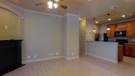 This single-floor space offers a clean, open layout designed for comfortable everyday living. The main living area is finished with light wood-style flooring that runs throughout, creating a bright and cohesive feel. Soft gray walls are accented by crisp white crown molding and matching baseboards, lending the space a polished, well-maintained character. A ceiling fan with a light fixture keeps the room comfortable and well-lit.

The living area flows naturally into the kitchen, which is partially separated by a structural column that adds architectural definition without closing off the space. The kitchen features a raised counter or bar-height surface, making it ideal for casual dining or entertaining. Pendant lighting hangs above the counter, providing focused task lighting with a subtle decorative touch. A stainless steel refrigerator is visible in the kitchen, hinting at modern appliance finishes throughout. Recessed lighting further illuminates the kitchen area, ensuring a bright and functional workspace.

Overall, the space presents a neutral, move-in-ready aesthetic with a harmonious blend of practical finishes and thoughtful design details, making it a versatile and inviting environment.