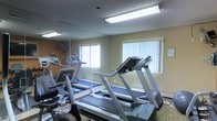 This single-level fitness center is purpose-built to support a range of cardiovascular workouts in a clean, functional environment. The room is equipped with multiple treadmills and a stationary bike, offering guests or members accessible options for daily exercise. A large wall-mounted mirror spans one side of the room, creating a sense of expanded space and allowing users to monitor their form during workouts. Natural light filters in through windows fitted with vertical blinds, complementing the overhead fluorescent lighting that keeps the space well-illuminated. The walls are finished in warm neutral tones, and a textured ceiling adds a classic touch to the interior. A ceiling fan assists with air circulation, contributing to a comfortable workout environment. The flooring features a patterned carpet, and the overall layout is efficiently arranged to maximize the use of the available floor space. Additional cardio equipment is visible toward the back of the room, rounding out the fitness offerings. The space is straightforward and practical, providing everything needed for a productive exercise session in a no-frills, welcoming setting.