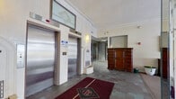 This single-floor commercial space offers a diverse layout across a generously sized footprint, combining formal reception areas with functional office and corridor spaces. The main lobby sets an elegant tone with stainless steel elevator doors, ornate crown molding, wall sconces, and decorative architectural details including a carved niche feature. Rich burgundy carpet runners accent the tiled flooring, while a substantial dark wood dresser adds a classic touch near the entry.

Adjacent to the lobby, a carpeted lounge area provides a welcoming atmosphere, highlighted by a crystal chandelier with a decorative ceiling medallion, crown molding, and large floor-to-ceiling windows with arched detailing that allow considerable natural light and views of an outdoor area. A fireplace unit, framed artwork, and leather seating contribute to the room's refined character.

The interior corridors extend toward glass-fronted office suites, identified by numbered signage, and feature drop ceilings with recessed fluorescent lighting and hard-surface flooring. Built-in mailbox banks line the hallways, indicating a multi-tenant environment. Some areas appear to be undergoing renovation, with construction materials visible behind glass partitions. Emergency exit signage and security cameras are present throughout, reinforcing the space's commercial functionality.