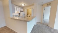 This single-story residence features an open-concept layout that seamlessly connects the kitchen and living area. The kitchen is outfitted with white appliances, including an electric range and a full-sized refrigerator, complemented by light-toned cabinetry that provides ample storage. An under-cabinet range hood adds functional ventilation above the cooktop. A peninsula counter extends from the kitchen, creating a natural division between the cooking space and the carpeted living area while offering additional workspace or casual seating. The neutral wall tones and flush-mount ceiling light keep the space feeling bright and cohesive. Adjacent to the kitchen, a dedicated laundry alcove houses a full-sized side-by-side washer and dryer, adding notable everyday convenience. The living area is finished with wall-to-wall carpet in a neutral tone, contributing to a comfortable, low-maintenance environment. A doorway visible from the main living space leads to additional rooms within the unit. Overall, the layout is practical and efficiently organized, making good use of the available floor plan across its single level.