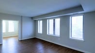 This single-floor space presents a clean, minimalist aesthetic characterized by neutral grey walls and dark hardwood-style flooring that grounds the room with a warm contrast. The main living area is notably open, offering a generous sense of space that feels uncluttered and versatile.

A standout feature of this room is the row of multiple windows along one wall, fitted with white horizontal blinds. These windows allow an abundance of natural light to fill the interior, creating a bright and airy atmosphere throughout the day. The windows also provide a visual connection to the outdoors, enhancing the overall sense of openness.

The ceiling features a subtle textured finish and is accented by a structural beam detail that adds a modest architectural element to the otherwise straightforward design. White baseboards trim the perimeter of the room, providing a crisp contrast against both the flooring and the walls.

Overall, the space is well-suited for flexible use, with its neutral palette and ample natural light making it adaptable to a variety of furnishing and décor styles. The combination of durable flooring, bright windows, and clean wall finishes gives the space a practical yet inviting character.