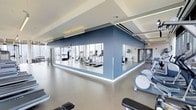 This contemporary fitness center is designed with both functionality and aesthetics in mind. The space features a clean, open layout with a notably high ceiling accented by recessed linear lighting that runs along the perimeter, providing consistent, even illumination throughout. A striking slate-blue accent wall serves as a central architectural element, housing a large mirrored section that visually expands the space and allows users to monitor their form during workouts.

Floor-to-ceiling windows line the perimeter, flooding the facility with abundant natural light and offering expansive city views, creating an energizing environment for exercise. The flooring is a smooth, neutral-toned surface suited for both cardio and strength training activities.

The equipment layout is thoughtfully arranged, with cardio machines including treadmills and ellipticals positioned to take advantage of the natural light and views. Additional strength and functional training equipment is visible throughout. The overall color palette — muted grays, white, and blue — reinforces a calm yet motivating atmosphere, making this fitness center a well-appointed facility for comprehensive workouts.