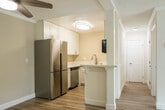 This single-floor unit at Olive Tree offers a clean, modern living environment defined by light-toned wood-style flooring that flows seamlessly throughout the space. The open living area is generously sized, bathed in natural light from a large sliding window dressed with vertical blinds, and fitted with a ceiling fan for year-round comfort. A textured ceiling adds subtle architectural character, while crisp white trim and neutral warm-toned walls create a bright, welcoming ambiance.

The kitchen is efficiently laid out in a U-shape configuration, featuring white shaker-style cabinetry with brushed nickel hardware, white quartz countertops, and a full suite of stainless steel appliances including a refrigerator, electric range, over-the-range microwave, and dishwasher. A pass-through opening connects the kitchen to the main living area, enhancing the sense of openness. A pendant light fixture illuminates the kitchen workspace.

The bedroom is a quiet, well-proportioned room with the same consistent flooring and a panel-style door, offering a simple and versatile layout. The bathroom is finished in a clean, all-white palette with a full tub and shower combo equipped with built-in shelving, chrome fixtures, wood-style flooring, and a vanity with ample counter space. Overall, this unit presents a well-maintained, move-in-ready space with cohesive finishes throughout.