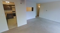 This single-floor residence offers a straightforward and functional layout designed for comfortable everyday living. The main living area is finished with neutral wall-to-wall carpeting, giving the space a soft, understated feel. White walls throughout create a bright, clean aesthetic that provides a versatile backdrop for any furnishing style. A defining architectural detail is the pass-through window cut into the partial wall that divides the living area from the kitchen, promoting an open flow and easy interaction between spaces. The kitchen, visible through the pass-through and accessible via an adjacent opening, features tile flooring that contrasts with the carpeted living area, and includes standard appliances such as a stove. A hallway branching off from the main living space leads to additional rooms, suggesting a private sleeping and bathroom area beyond. Interior lighting is provided by ceiling-mounted fixtures. The overall layout is compact yet practical, with clearly defined zones for living, cooking, and private use. The neutral palette and simple finishes make this a move-in-ready space that is easy to personalize.