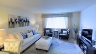 This single-floor space showcases a thoughtfully designed living area centered around a cohesive blue-and-white color palette. The living room serves as the focal point, featuring a plush white sofa adorned with blue floral accent pillows, paired with two tufted dark navy armchairs arranged symmetrically around a round side table. A large rectangular ottoman in crisp white doubles as a coffee table, adding both functionality and visual balance to the seating arrangement.

A generously sized window dressed with layered window treatments — including sheer white vertical blinds and a pleated fabric valance in soft gray-blue tones — allows abundant natural light to fill the room while maintaining a sense of privacy. The light walls and smooth white ceiling enhance the bright, airy feel of the space.

Underfoot, a decorative area rug with a subtle botanical pattern in blue and beige anchors the seating arrangement. Wall décor includes a series of blue floral canvas prints, reinforcing the space's elegant, coordinated aesthetic. A floor lamp and decorative accents complete the polished, well-curated look of this inviting living area.