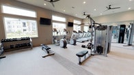 This modern fitness center is designed to support a comprehensive workout experience in a bright, open environment. The space is outfitted with a row of cardio machines, including treadmills and ellipticals, positioned along a wall of large horizontal windows that flood the room with natural light and provide views of the surrounding area. Ceiling fans are installed overhead to maintain airflow and comfort during workouts. Wall-mounted flat-screen TVs are strategically placed throughout the space, allowing users to stay entertained while exercising. A Precor cable machine anchors the strength training area, accompanied by adjustable workout benches for functional and free-weight exercises. The flooring is light-toned and durable, suited for high-traffic fitness use. Recessed lighting complements the natural light, ensuring the space remains well-lit at all hours. The neutral color palette of soft grays and whites gives the facility a clean, contemporary aesthetic. A large mirror on one wall enhances the sense of openness while supporting form-checking during strength exercises. Overall, the fitness center presents a professional and inviting atmosphere for residents or guests pursuing their health and wellness goals.