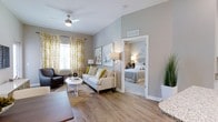 This well-appointed 2-bedroom, 2-bathroom residence offers a thoughtfully designed single-floor layout that maximizes both comfort and functionality. The open-concept living area serves as the heart of the home, featuring warm hardwood-style flooring that flows seamlessly throughout the space. Vaulted ceilings with a ceiling fan add a sense of airiness and volume to the living room, which is bathed in natural light through large windows dressed with stylish patterned curtains in a neutral and gold palette. The living area provides ample room for relaxed seating arrangements, complemented by tasteful wall art and modern lighting fixtures. A clear sightline connects the living space directly to the bedrooms, one of which is visible through a wide doorway and showcases plush bedding and soft, inviting tones. The interior color palette is kept neutral with soft gray walls, creating a calm and cohesive ambiance throughout. Granite countertop accents are visible at the edge of the kitchen area, hinting at quality finishes carried through the home. The overall design balances contemporary style with everyday livability, making this a functional and attractive residence.