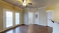 This single-floor residence presents a clean, well-appointed interior that balances comfort and practicality. The main living area is anchored by rich, dark hardwood floors that extend throughout, lending warmth and a polished look to the space. Crown molding traces the upper perimeter of the room, adding a classic architectural detail that elevates the overall aesthetic. A ceiling fan with an integrated light fixture is centrally positioned, ensuring efficient air circulation and overhead illumination. The walls are finished in a neutral beige tone, creating a versatile backdrop that complements a variety of interior styles. Natural light enters generously through a full-length glass door fitted with adjustable white blinds, offering both brightness and privacy control. Multiple interior doorways provide access to additional areas of the home, with one opening revealing a well-lit adjacent room. Visible utility features, including a ventilation panel and wall-mounted controls, reflect a thoughtfully functional design. White trim and baseboards throughout provide a crisp contrast to the warm flooring and walls, tying the space together with a cohesive and tidy finish. The overall ambiance is bright, open, and move-in ready.