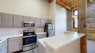 Located at 560 Mart St in Muskegon, Michigan, Unit 102 is a single-floor loft-style residence that masterfully combines industrial heritage with contemporary design. The space is defined by striking exposed brick walls and warm wood-beam ceilings, lending a sense of history and character throughout. Natural light pours in through large, wood-framed multi-pane windows, illuminating the interiors and highlighting the textural contrast between the rustic brickwork and clean, modern finishes. The kitchen is a standout feature, outfitted with sleek, gray wood-grain cabinetry extending to impressive heights, stainless steel appliances including a range, microwave, and refrigerator, and bright white countertops that include a central island providing additional workspace. Wood-look luxury vinyl flooring runs through the kitchen and living areas, adding warmth underfoot. The bedrooms feature plush carpet and expansive brick accent walls paired with generously sized windows that invite the outdoors in. The bathrooms are finished with modern floating vanities, white countertops, full tub-shower combinations, and wood-look tile flooring, offering a clean and functional aesthetic. Throughout the unit, soaring ceiling heights reinforce the open, airy loft ambiance that makes this Muskegon residence truly distinctive.