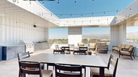 This sophisticated rooftop amenity space seamlessly blends indoor and outdoor living across a single, well-appointed level. The interior lounge is defined by its distinctive coffered ceilings painted in a cool slate blue, warm light wood flooring, and large sliding glass doors that connect directly to the outdoor terrace. A statement dark marble island anchors the interior, suspended beneath a series of globe pendant lights on brushed gold hardware. Dark walnut cabinetry lines the kitchen wall alongside sleek white countertops, a built-in beverage refrigerator, and tall narrow windows that draw in natural light. The lounge area features an olive green velvet sectional, a patterned teal and white area rug, and a bold sputnik chandelier, creating an inviting and eclectic gathering space. Black-framed glass doors add an architectural accent that separates zones while maintaining visual flow throughout. Outside, the expansive terrace offers multiple dining and lounge configurations, a built-in outdoor kitchen with a gas grill, and string lights overhead. Comfortable seating areas frame panoramic views of a mountain range and surrounding cityscape, making this rooftop an ideal setting for both casual relaxation and entertaining.