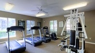 This fitness center offers a practical and comfortable environment for exercise, thoughtfully designed to accommodate a variety of workout needs. The space is outfitted with multiple treadmills and cardio machines, providing ample options for cardiovascular training. A multi-station strength training unit adds versatility, allowing for a range of resistance exercises within the same room. The flooring features wall-to-wall carpet, lending a clean and tidy appearance throughout. Neutral-toned walls create a calm, focused atmosphere, while a ceiling fan with an integrated light fixture helps maintain comfortable air circulation. Several windows along the walls invite natural light to fill the space, offering pleasant views of the surrounding greenery outside. Framed posters and informational displays are mounted on the walls, adding motivational and instructional elements to the environment. Flush-mounted fluorescent lighting ensures the room remains well-lit at all times. The overall layout is open and organized, making efficient use of the available floor space while maintaining clear pathways between equipment. This fitness center is a functional and inviting facility suited for daily exercise.