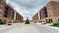 This single-floor residence is situated within a striking multi-building complex that blends traditional brick masonry with modern architectural elements, including contrasting light and dark façades and clean contemporary lines. The complex is thoughtfully designed with a welcoming streetscape, featuring a central landscaped roundabout with manicured shrubs, ornamental trees, and a classic lamp post that anchors the entrance corridor. Private balconies are featured throughout the building exterior, offering residents outdoor space and views of the surrounding area. The grounds are well-maintained, with neatly trimmed hedgerows lining the walkways and drive, contributing to a polished and cohesive community aesthetic. Stone and brick detailing at the building base adds a sense of permanence and quality craftsmanship to the overall design. The layout of the complex creates a sense of arrival, with symmetrical building placement framing a central approach that feels both organized and inviting. The surrounding environment suggests a suburban setting with easy access to parking and pedestrian pathways. Overall, the space reflects a modern residential community that balances architectural character with practical amenities and thoughtfully designed outdoor common areas.