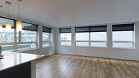 This single-floor space offers a clean, modern aesthetic defined by an open-plan layout and an abundance of natural light. The main living area features warm-toned hardwood-style flooring that runs continuously throughout, providing a cohesive and inviting foundation. Crisp white walls and a smooth white ceiling enhance the sense of openness and brightness.

A standout feature of this space is its generous window arrangement, with multiple large windows lining two walls. These windows are fitted with sleek black roller blinds, offering both light control and privacy while maintaining a contemporary look. The windows provide views of the surrounding urban streetscape, connecting the interior to its city environment.

The layout is notably open, with no visible partitions dividing the main living area, allowing for flexible furniture arrangement and a spacious feel. A partial countertop or surface is visible at the edge of the frame, suggesting proximity to a kitchen or breakfast bar area. Overall, the space presents a minimalist, move-in-ready environment that balances functionality with a fresh, modern design sensibility suited to urban living.