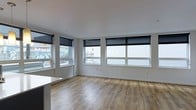 This single-floor space offers a clean, modern aesthetic defined by an open-plan layout and an abundance of natural light. The main living area features warm-toned hardwood-style flooring that runs continuously throughout, providing a cohesive and inviting foundation. Crisp white walls and a smooth white ceiling enhance the sense of openness and brightness.

A standout feature of this space is its generous window arrangement, with multiple large windows lining two walls. These windows are fitted with sleek black roller blinds, offering both light control and privacy while maintaining a contemporary look. The windows provide views of the surrounding urban streetscape, connecting the interior to its city environment.

The layout is notably open, with no visible partitions dividing the main living area, allowing for flexible furniture arrangement and a spacious feel. A partial countertop or surface is visible at the edge of the frame, suggesting proximity to a kitchen or breakfast bar area. Overall, the space presents a minimalist, move-in-ready environment that balances functionality with a fresh, modern design sensibility suited to urban living.