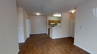 This single-floor space features an open-concept layout that seamlessly connects the living area to the kitchen, creating a functional and cohesive environment. Rich, dark hardwood-style flooring extends throughout the main living space, adding warmth and visual continuity to the interior.

The kitchen is positioned centrally and defined by a half-wall pass-through that opens to the living area. Warm-toned wood cabinetry lines the kitchen walls, complemented by white appliances including a refrigerator and range. The overhead lighting in the kitchen provides bright, focused illumination, and ample counter space supports everyday meal preparation.

The living area is generously sized with neutral-toned walls that contribute to a clean, understated aesthetic. Flush-mount ceiling lights provide even lighting throughout the space. Multiple doorways are visible from the main living area — one leading to what appears to be a closet with bifold doors, and another to an additional room, suggesting practical storage and flexible room configurations.

Overall, the space offers a straightforward, functional layout with consistent finishes, making it well-suited for everyday living.