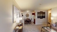 Located at Brookwood at Ridge in Ridge, NY, this single-floor residence is thoughtfully laid out to maximize comfort and functionality. The living room serves as a welcoming centerpiece, featuring plush wall-to-wall carpeting, a large window with sheer vertical blinds that invite natural light, and neutral-toned walls that create a calm, versatile backdrop. A decorative mirror and soft lighting add warmth to the space.

Adjacent to the living room, the dining area offers a cozy setting for meals, complete with carpeted flooring, a stylish wall clock as a focal point, and convenient access to a closet for additional storage. The kitchen is efficiently designed with rich cherry wood cabinetry, granite-style countertops, hardwood-style flooring, and white appliances including a range and refrigerator. A decorative oval mirror adds a charming detail to the kitchen walls.

The bedroom is a restful retreat featuring carpeted floors, a window dressed with sheer curtains providing soft natural light, and a sliding closet offering ample storage. The full bathroom is finished with tile flooring, a rich wood vanity with an integrated sink, a medicine cabinet mirror, and a shower-tub combination. Overall, the residence presents a clean, comfortable, and practical living environment.