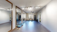 This single-room fitness and yoga studio offers a thoughtfully designed space for a range of exercise activities. The elongated layout provides an open, unobstructed floor area finished with smooth, light-colored flooring that transitions to wood-tone planks near the entry — ideal for both high-energy workouts and mindful movement practices.

A defining feature of the studio is the full wall of large, wood-framed mirrors along one side, which visually expand the space while allowing users to monitor their form. Overhead, a combination of adjustable track lighting and ceiling fans ensures the room remains well-lit and comfortable during use. Natural light enters through a row of windows along the far wall, offering views of outdoor greenery and creating a bright, inviting atmosphere.

The studio accommodates spin bikes positioned on protective floor mats, TRX suspension straps mounted to the walls, and step platforms and yoga mats for group fitness or individual sessions. A dedicated entry area near the wood-framed glass door features a built-in cubby storage unit stocked with small equipment such as dumbbells, resistance bands, and yoga blocks — keeping the main floor clear and functional. The overall ambiance is clean, purposeful, and well-organized.