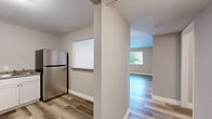 This thoughtfully designed single-floor residence offers a complete layout comprising a living room, kitchen, bedroom, bathroom, and hallway, all unified by durable wood-look plank flooring and a neutral, contemporary color palette throughout. The kitchen is equipped with crisp white shaker-style cabinetry, granite-look countertops with an L-shaped configuration, a double stainless steel sink, and a stainless steel refrigerator — providing ample storage and prep space for everyday cooking. A pass-through window connects the kitchen to the adjacent living area, enhancing the open feel of the space. The living room is bright and inviting, featuring a large window that frames views of lush greenery, allowing generous natural light to fill the room. The bedroom is a comfortable, well-proportioned space with its own window overlooking the outdoors, complemented by the same wood-look flooring and clean wall finishes found throughout the unit. The hallway connects the main living spaces efficiently and includes a sliding-door closet, offering convenient storage. The bathroom features a well-lit vanity with a lighted mirror, chrome fixtures, and a clean, modern aesthetic. Consistent white baseboards and neutral-toned walls tie the entire residence together, creating a cohesive, move-in-ready environment.