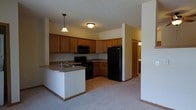 Located at 1910 Hawks Ridge Dr in Verona, Wisconsin, this single-floor residence offers a practical and comfortable layout designed for everyday living. The heart of the home is an open-concept kitchen that flows seamlessly into the living area, creating a spacious and connected feel. The kitchen is outfitted with warm honey-toned wood cabinetry that provides generous storage, complemented by black appliances including a refrigerator, electric range, and over-the-range microwave. A peninsula with an integrated sink and pendant lighting above serves as both a functional workspace and a casual gathering spot, bridging the kitchen and living areas. The flooring transitions from resilient vinyl in the kitchen to plush carpeting in the adjoining living space, adding warmth underfoot. Ceiling-mounted lighting keeps the space well-illuminated throughout. A hallway visible from the main living area leads to additional rooms, suggesting a thoughtful floor plan that balances open communal spaces with private retreats. The overall ambiance is warm and inviting, with a neutral color palette that provides a versatile backdrop for a variety of décor styles.