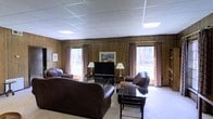 Located at 5640 Keele St in Jackson, Mississippi, this single-story space offers a functional and inviting layout designed for comfort and ease of use. The main living area serves as the centerpiece of the space, showcasing rich wood-paneled walls that lend a warm, classic character throughout. A drop-panel ceiling with recessed lighting ensures consistent, well-distributed illumination across the room. Large windows dressed with patterned curtains allow natural light to filter in while framing views of the surrounding exterior, contributing to an open and airy feel despite the cozy aesthetic. The seating area is generously appointed with plush leather sofas and accent chairs arranged around a glass-top coffee table, creating an ideal setting for relaxation or social gatherings. A flat-screen television is centrally positioned along the main wall, flanked by traditional table lamps that add a warm, ambient glow. Light-toned tile flooring ties the space together, offering both durability and a clean contrast to the darker wood wall paneling. The overall ambiance is welcoming and functional, blending traditional design elements with comfortable, practical furnishings suited for everyday use.