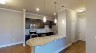 This single-floor unit offers a thoughtfully designed open-concept layout that seamlessly connects the kitchen and living areas. The kitchen is a standout feature, equipped with dark espresso cabinetry, granite countertops, a subway tile backsplash, and a full suite of stainless steel appliances including a range, microwave, dishwasher, and refrigerator. A curved granite-topped peninsula with pendant lighting above serves as both a functional workspace and a natural divider between the kitchen and the expansive living area. Crown molding throughout adds a refined architectural detail, while warm hardwood-style flooring flows consistently across the main living spaces. The living area is generously proportioned, offering flexible arrangements and a bright, open ambiance. The bedroom features plush carpeting and a built-in closet with sliding doors and multi-level shelving for ample storage. Two bathrooms are included: one with a soaking tub and tiled shower surround with accent detailing, dark cabinetry, and granite vanity; and a second elongated bathroom with a walk-in style shower, granite vanity, and additional upper cabinetry. Throughout, neutral gray walls, recessed lighting, and clean white trim create a cohesive, contemporary aesthetic.
