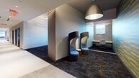 The Six Points Bloomfield Workspace is a thoughtfully designed single-floor professional environment in Bloomfield, New Jersey, offering a variety of work settings to accommodate different working styles. The layout balances open circulation areas with dedicated private and semi-private zones, creating a versatile and functional atmosphere.

The common corridor features polished terrazzo flooring that transitions to dark patterned carpet, establishing clear zones throughout the space. Distinctive high-back pod chairs provide semi-private seating areas ideal for focused individual work or informal conversations, positioned alongside glass-walled private offices that allow visual connectivity while maintaining separation.

A formal conference room is outfitted with a spacious white table, ample seating, a wall-mounted display screen, and striking geometric pendant lighting fixtures suspended from the ceiling, lending the room a contemporary, design-forward character. Warm wood paneling accents the room's rear wall, adding depth and sophistication.

A dedicated focus area features built-in wood-framed alcove booths with deep-toned interior walls, individual task surfaces, and upholstered bench seating—ideal for heads-down work. Natural light enters through windows overlooking the surrounding Bloomfield streetscape.

An amenity room finished with geometric patterned tile walls in gold and grey tones includes shower facilities and vanity areas with warm wood cabinetry, offering convenient on-site wellness accommodations for building occupants.
