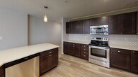Located at 1120 Euclid Ave in Cleveland's vibrant Euclid Corridor, this well-appointed unit offers a thoughtfully designed single-floor layout with modern finishes throughout. The open-concept living and kitchen area serves as the heart of the space, featuring light wood-style flooring, recessed lighting, and a curved island with a built-in sink and sleek white countertop, illuminated by stylish pendant lights. The kitchen is outfitted with rich dark-wood cabinetry, a decorative arabesque tile backsplash, stainless steel appliances including a range and built-in microwave, and generous counter space along an L-shaped configuration. Large, black-framed windows in the living area flood the space with natural light and frame views of the surrounding urban streetscape. The bedrooms are finished with plush wall-to-wall carpeting and neutral-toned walls, providing a calm and comfortable retreat. One bedroom features a wide frosted-glass window panel. The full bathroom includes a soaking tub with marble-style tile surround, a dark-wood vanity with undermount sink, and wood-look flooring. A second bathroom offers a vanity with matching cabinetry and additional storage. The overall aesthetic is clean, contemporary, and move-in ready.