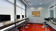 This single-floor space is designed as a functional and modern computer or business center, offering a clean and organized environment for productivity. A row of iMac desktop computers lines a sleek, dark wall-mounted desk that runs along the large windows, allowing natural light to filter in through roller shades. Adjustable bar-height stools with black cushioned seats and polished steel bases are positioned at each workstation, providing comfortable seating at counter height. The bold red flooring creates a striking contrast against the light gray walls, giving the space a contemporary and energetic feel. Recessed ceiling lights supplement the natural light, ensuring the area remains bright and well-illuminated. A cork bulletin board mounted on the far wall serves as a practical information hub for notices and announcements. A drop ceiling with ventilation grilles maintains a clean, organized overhead aesthetic. The overall ambiance is professional yet approachable, making the space well-suited for work, research, or collaborative use. The open layout allows for ease of movement throughout the room.