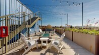 Located at 521 W Colorado St in Glendale, California, this rooftop terrace offers a well-appointed outdoor retreat with panoramic views of the surrounding Glendale cityscape and the San Gabriel Mountains beyond. The terrace is finished with smooth concrete pavers and bordered by a row of large raised planters filled with lush succulents, flowering plants, and greenery, adding a natural, garden-like character to the elevated setting.

At the heart of the space is a built-in fire pit with a glass-bead bed, surrounded by white Adirondack chairs and a side table, creating an inviting lounge area ideal for gathering. A wicker pod daybed provides an additional seating option, offering a cozy nook with cushioned comfort and open-air views. Overhead, strands of globe string lights are strung across the terrace, lending a warm ambiance suited for evening use.

The terrace is set against a bold blue corrugated metal façade, with a metal staircase providing access to an upper level. The industrial architectural elements contrast effectively with the relaxed, resort-style furnishings. The open layout provides generous space for both lounging and social gathering, making this rooftop a functional and visually striking amenity with commanding views under the Southern California sky.